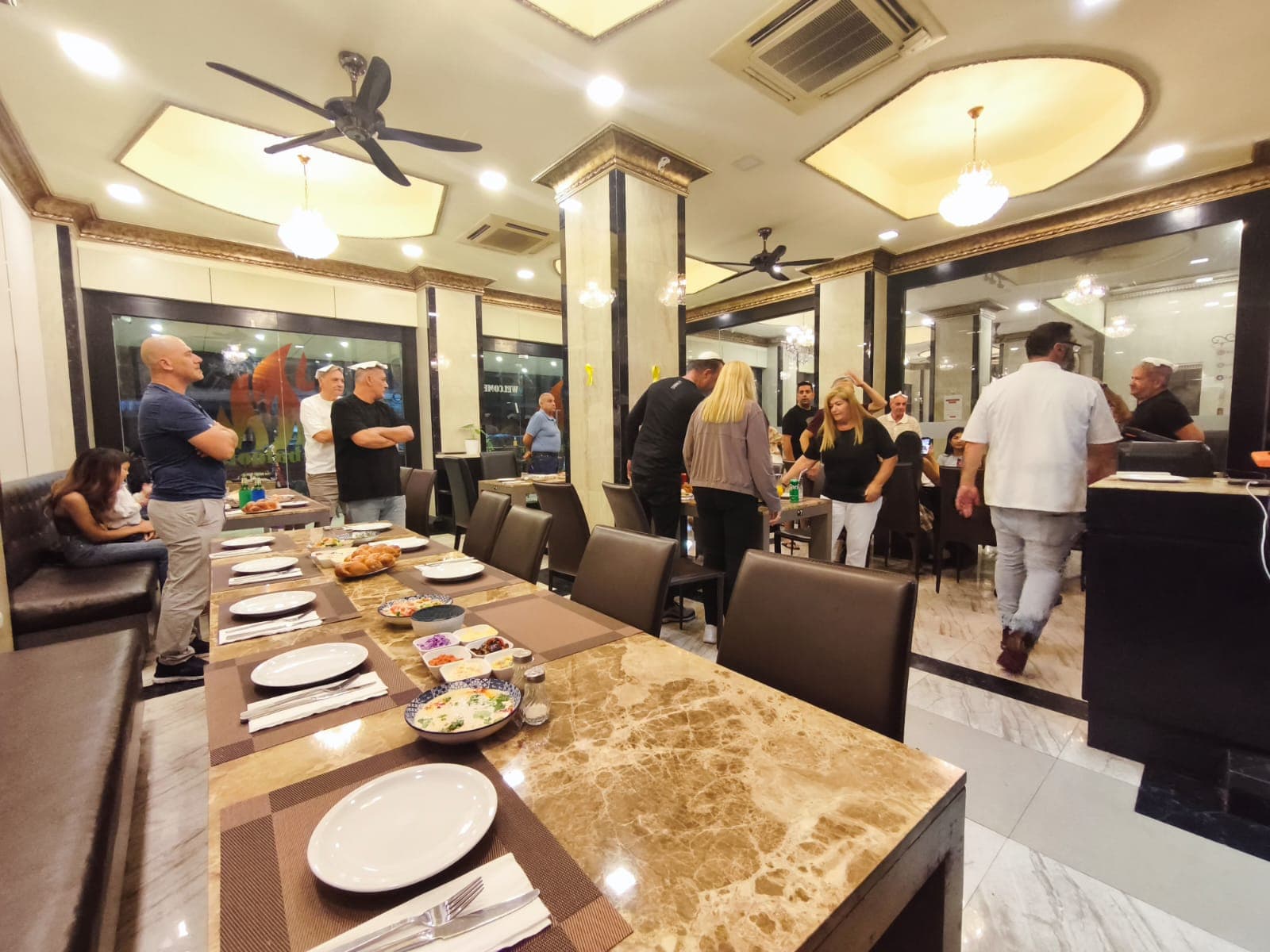 People mingling in an ornate restaurant with marble tables set with salads and challah bread.